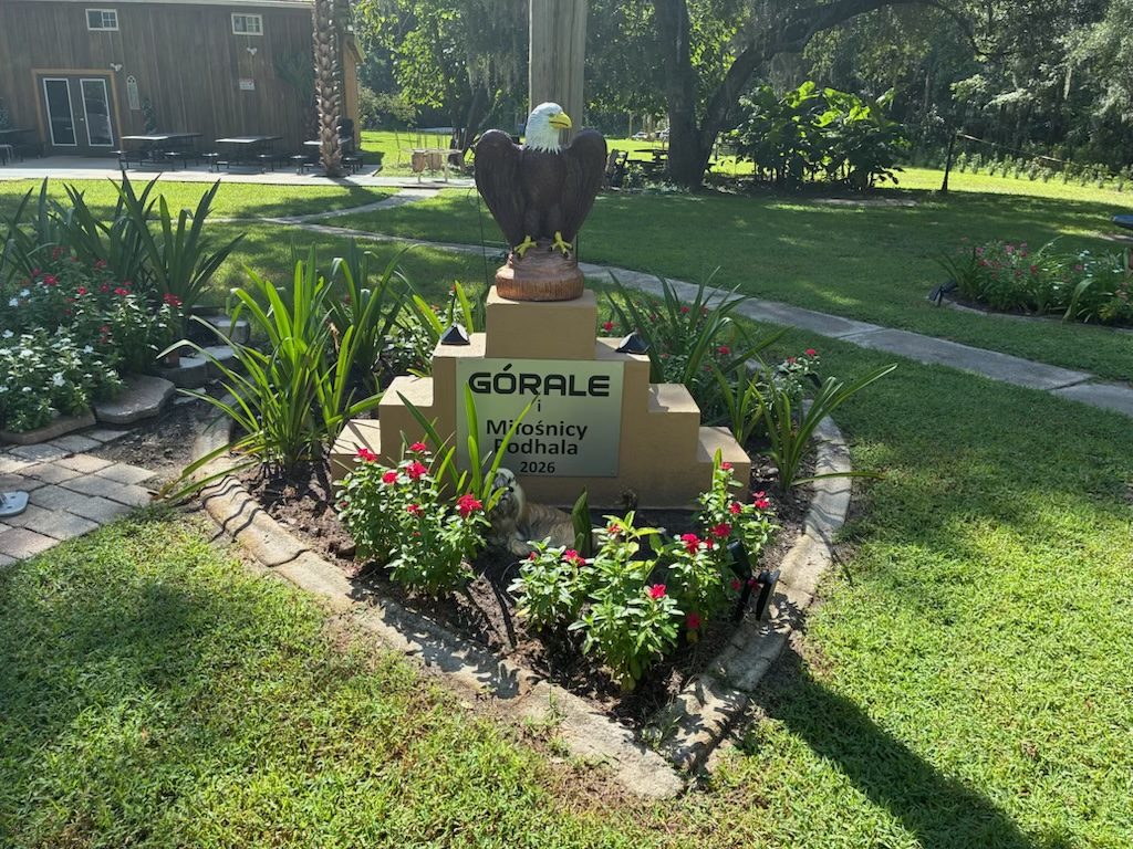 A stone monument on the farm topped with a carved eagle, inscribed Górale i Miłośnicy Podhala 2026, surrounded by red flowers.